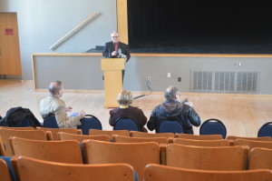 Three people sitting in a lecture hall looking at a man on stage reading from a book.