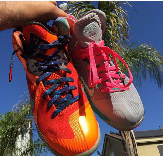 A hand holding a pair of mismatched Nike shoes. The left shoe is orange with blue laces and the right side is grey with pink laces.