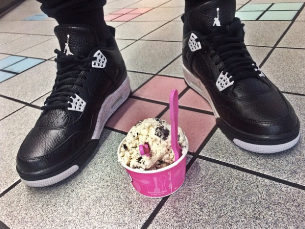 A scoop of Baskins Robbins cookies and cream ice cream with two spoons in it sitting on the ground in between a pair of black and white shoes on someones feet.