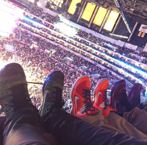 The legs of three people wearing sneakers watching a basketball game inside a stadium/arena.