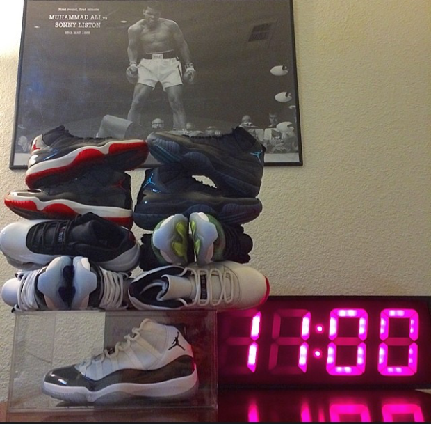 A shoe in a see through display box with shoes piled on top of it. To the right of the box is a digital clock that reads 11:00. In the background hangs a picture of boxer Muhammad Ali in the ring looking down at opponent Sonny Liston.