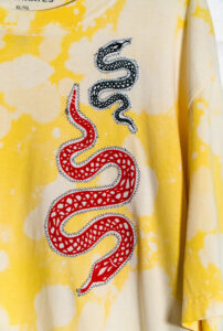 A close-up of a yellow tie-dye shirt. Two snakes are embroidered onto the shirt, one is red and large and the other is blue and small.