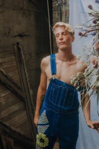A blonde person with short blonde hair stands outside beside what looks to be a shed. They are wearing dark blue denim overalls over top of bare skin, and the overalls have an embroidered yellow flower on the left leg, and two triangular pieces of fabric with floral designs on it.  There are flowers peeking out in the frame, slightly over the persons body. There is a white sheet in the background covering about half of the frame.