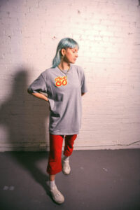 A white person with blue hair stands with their hand on their back, in a pose. They are inside, against a white brick wall. They are wearing a large grey T-shirt with the logo "GAY," on it. They are wearing red pants and white shoes.