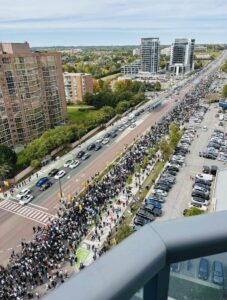 Photo of hundreds of protestors filling the streets in Toronto. The photo is taken from up high, from a balcony. It is daytime and sunny.