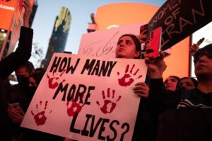 Protestor stands in a crowd holding a sign reading "How many more lives?" With red handprints painted on. The sun seems to be setting.