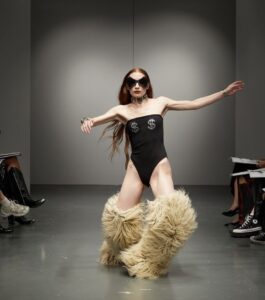 A white model with long orange hair is in mid-trip walking down the AVAVAV runway. Their arms are out long for balance and they're wearing a sleeveless black bodysuit with two silver dollar signs on each breast. They're wearing large over-the-knee ultra fluffy boots. They're also wearing a choker. The entire background is grey.