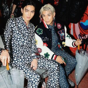 Two models sit side by side holding clear umbrellas by their sides. One model has short blonde hair, one model has short black hair. The models both appear male and are wearing pieces from The Tommy Factory. The model with black hair is wearing a matching black and white pattern suit with flowers, and the other model is wearing the same suit but in blue, and overtop he is wearing a sports jacket with patches.