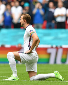 Harry Kane kneels on one knee on the pitch with his hands on his hips. He wears the Pride armband.