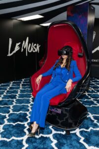 A woman in a blue suit sits on a Voyager chair, a black chair with red velvet interior. She is wearing a VR headset.