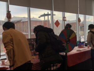 Two people are checking out the lucky raffle booth, next to the spin the wheel. There are stickers and decorations on the window. The window is a see-through glass through which we can see the lakeshore buildings. There is another student with a cap at the next station and there is an organizer behind that station